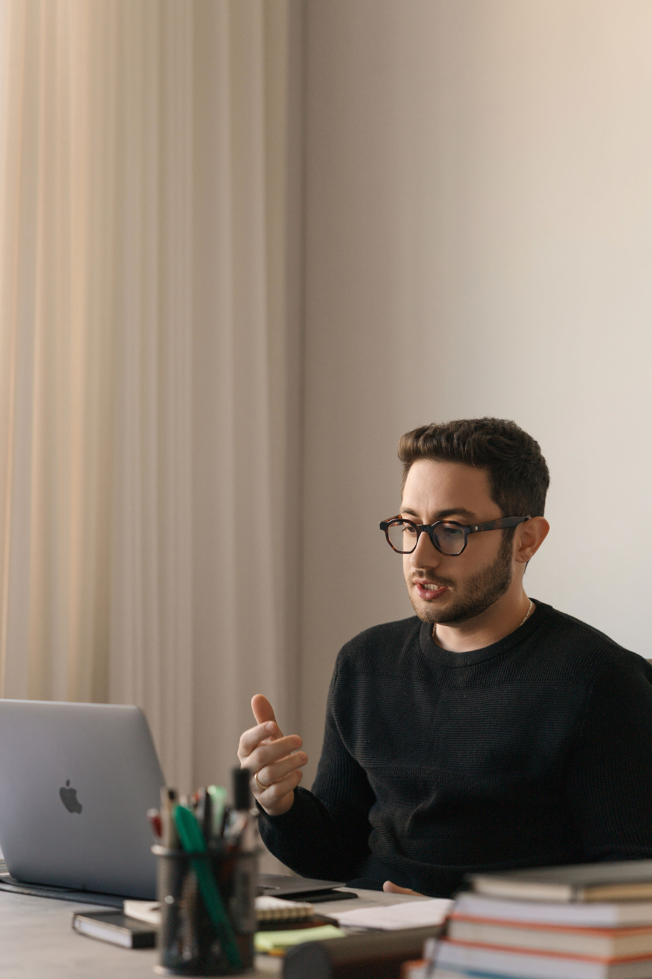 Lucas Vincci conversando à mesa de trabalho, de óculos e suéter preto, com o notebook ao lado